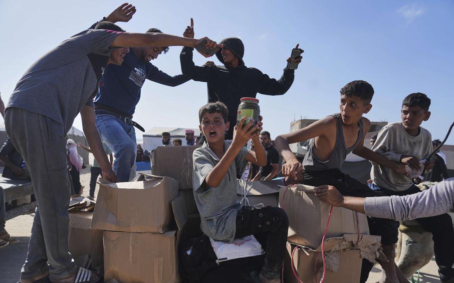 A Palestinian boy holds up a jar while sitting among boxes of food and humanitarian aid and other Palestinians stand around him.