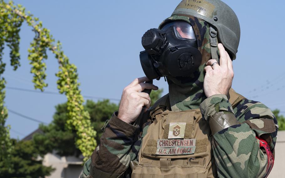 A U.S. Air Force service member wearing camouflage uniform and a military helmet puts on a black gas mask during a training exercise. The name tape “CHRESTENSEN” and “U.S. AIR FORCE” are visible on the uniform, along with military rank insignia. The background shows green foliage and a clear blue sky.
