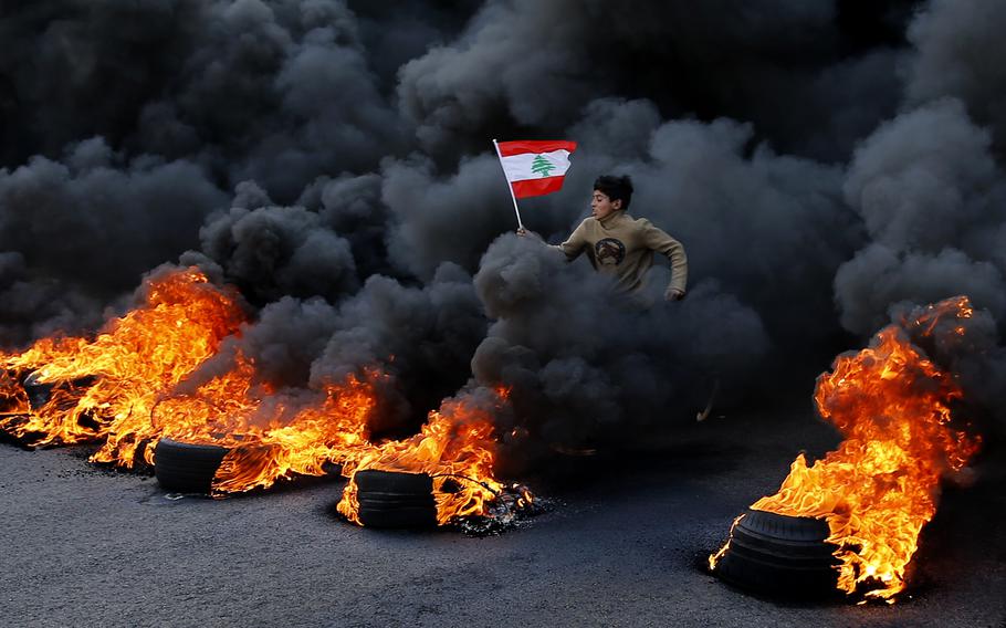 An anti-government demonstrator holds a national flag and runs across tires that were set on fire to block a main highway during a protest against a ruling elite they say has failed to address the economy's downward spiral, in the town of Jal el-Dib, north of Beirut, Lebanon, Tuesday, Jan. 14, 2020. Lebanon and Sri Lanka may be a world apart, but they share a history of political turmoil and violence that led to the collapse of once-prosperous economies bedeviled by corruption, patronage, nepotism and incompetence.