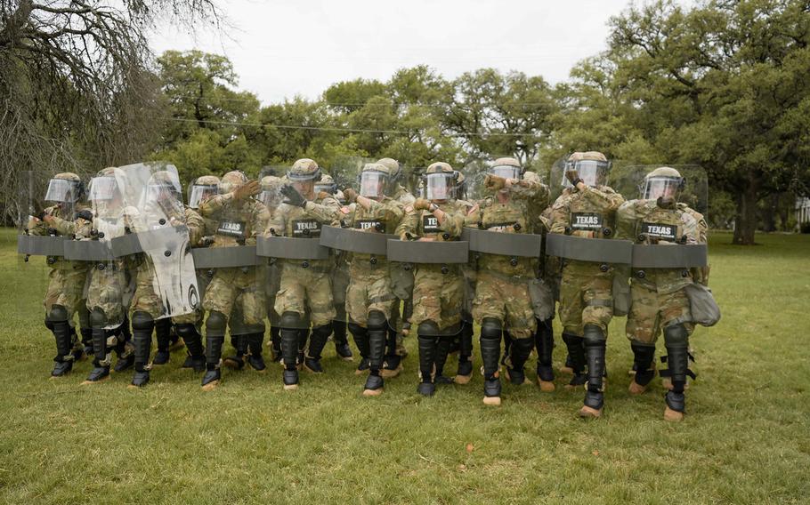National Guard troops wearing helmets with face guards and holding shields march through an empty field.