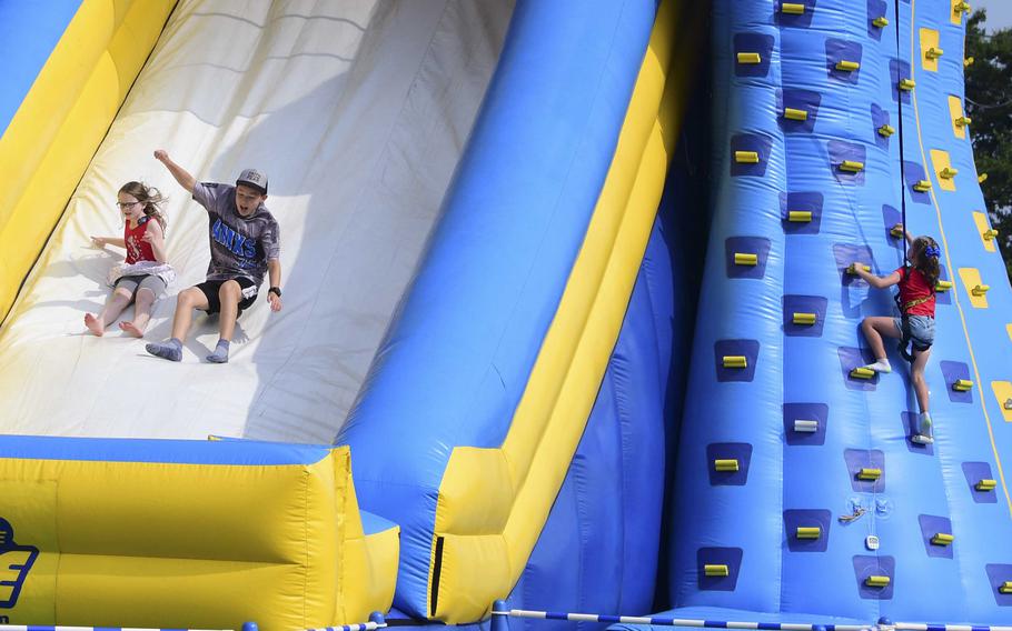 Two children travel down a large inflatable slide on the left of the frame, while a child in a harness climbs up the hand-holds of an inflatable climbing wall on the right side of the frame.