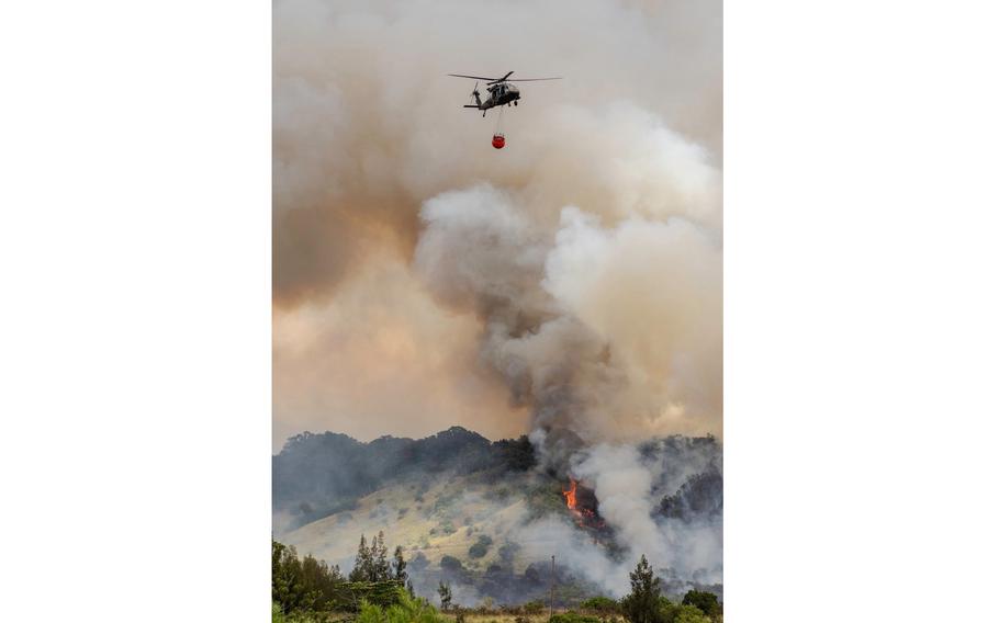 A large plume of smoke billows into the sky from a fire in a forested area as a helicopter hovers above to drop water from a red basket.