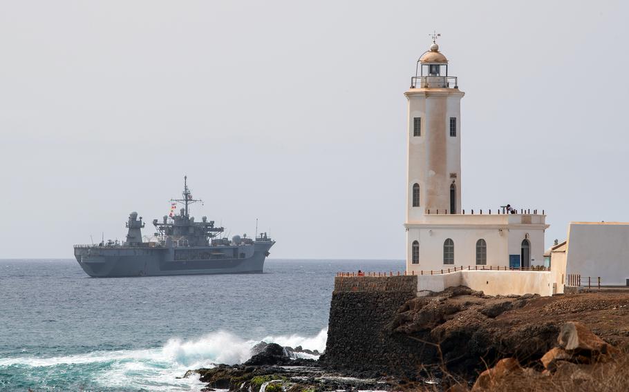 The USS Mount Whitney sails in the distance past a lighthouse.