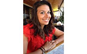A woman with long dark hair and wearing a bright red ruffled top, smiles while seated at a restaurant table with a menu in front of her.