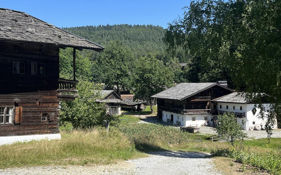 Traditional Alpine village scene showing historic wooden and white-painted buildings with characteristic balconies and steep roofs, surrounded by lush green hills and forest.