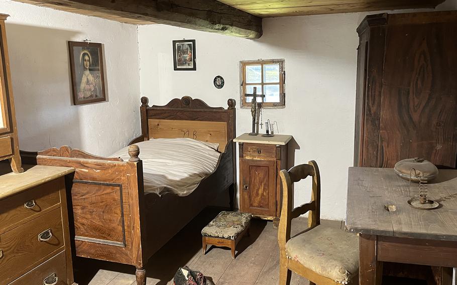 Interior of a historic bedroom featuring a wooden bed with carved headboard, traditional furniture, religious artwork on white walls, exposed wooden ceiling beams, and period furnishings.