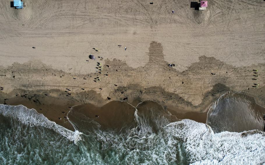 This aerial photo taken with a drone shows beachgoers as workers in protective suits continue to clean the contaminated beach in Huntington Beach, Calif., on Oct. 11, 2021. Officials were investigating an oil sheen spotted Saturday, Nov. 20, near last month’s crude pipeline leak off Southern California’s coast. The U.S. Coast Guard said in a statement the oil sheen is about 70 feet by 30 feet and that it has dispatched “pollution responders, aircraft and boats” to investigate.