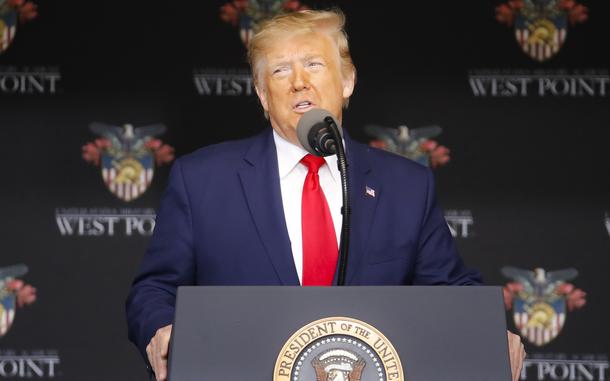 Donald Trump stands at a podium and speaks into a microphone in front of a dark curtain reading “West Point” and displaying the emblem of the United States Military Academy 