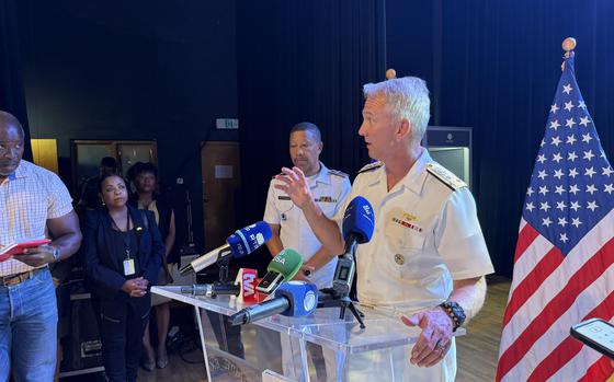 A man stands at a clear podium and talks into several mics while reporters stand nearby taking notes.