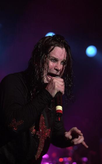 A vertical photo of Ozzy Osbourne singing into a microphone while making the rock and roll gesture with his other hand.