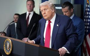 Donald Trump, wearing a dark suit and red tie, stands at a podium with a microphone as other officials in suits stand behind him.