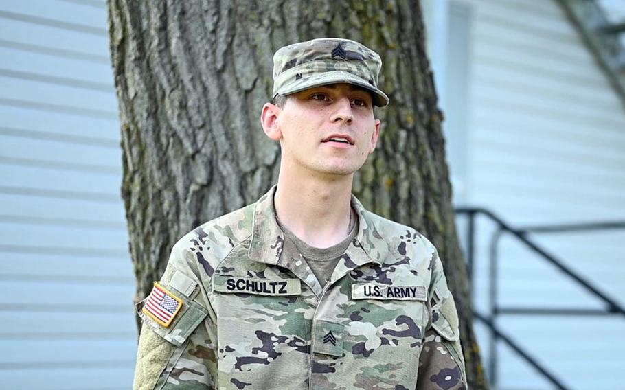 A U.S. Army soldier in camouflage uniform and cap, identified as Sergeant Schultz, stands outdoors in front of a tree and a white building.