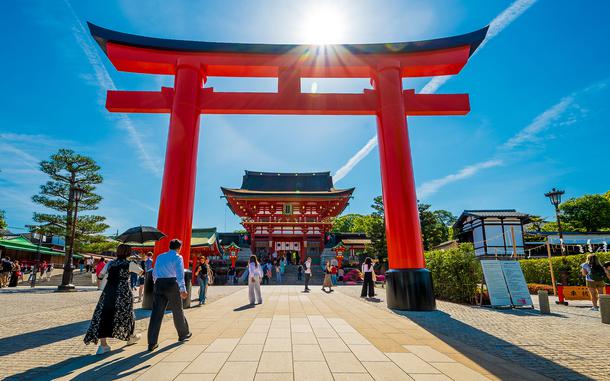 Tourists walk on a plaza around a large red Japanese torii gate, seen from ground level, with the sun shining above the gate and a temple in the background.