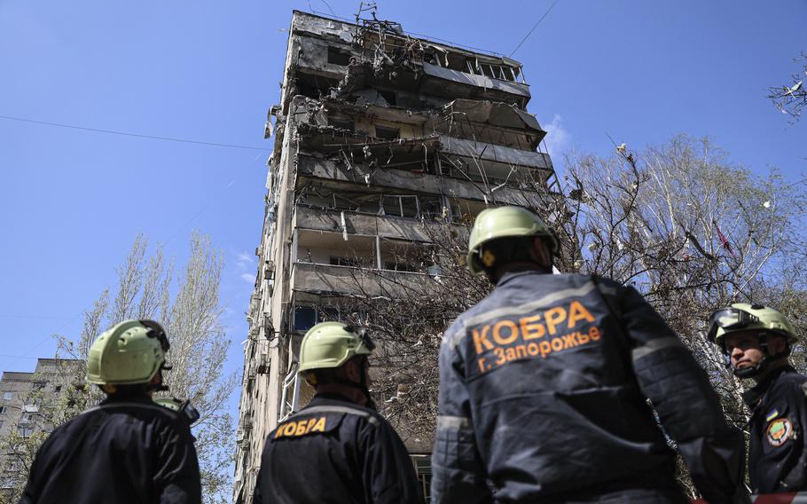 Rescue workers in hard hats look up a a damaged multi-story building.