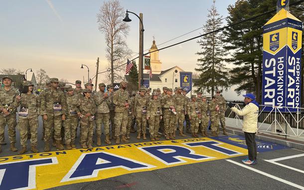 Uniformed troops stand  a listen to Boston Marathon race director Dave McGillivray behind  writing on the ground that says “Boston Marathon 2025 Start.”