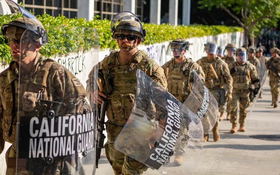 National Guard members carrying shields walk past a wall covered in graffiti. 
