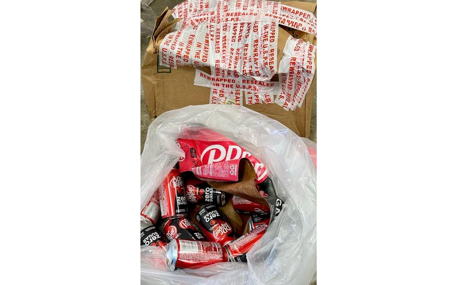 A garage bag filled with soda cans of Dr. Pepper, seen from above, sits next to a dented cardboard box with white and red tape.