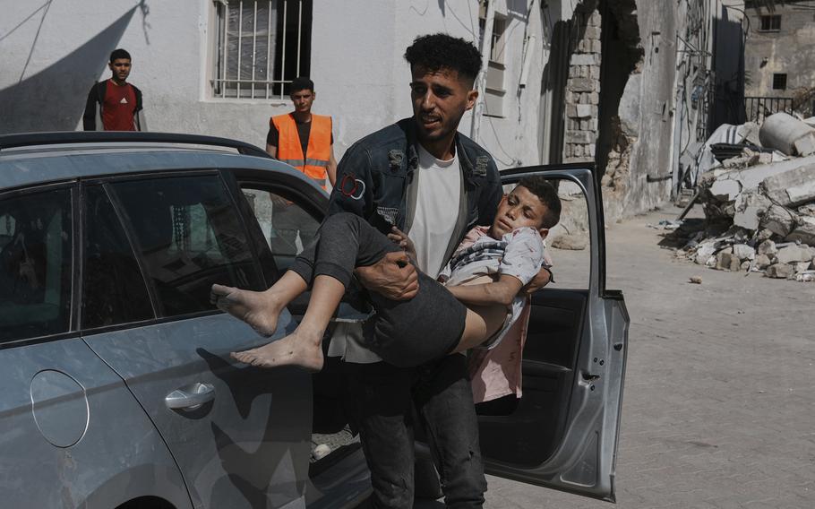 A Palestinian man carries an injured boy from a car with onlookers and a damaged building in the background.