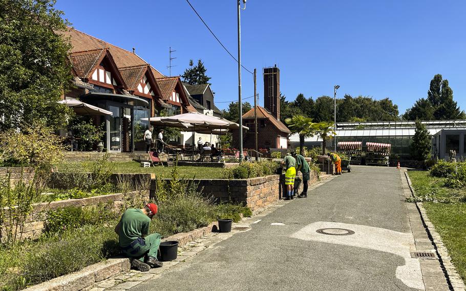 Workers in green uniforms maintain landscaping along a residential street featuring traditional Tudor-style houses with red tile roofs and timber framing. The scene shows several maintenance workers tending to planted areas bordered by brick retaining walls, with outdoor dining furniture visible on a terrace in the background. 