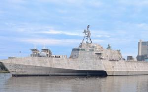 A gray-hulled military ship sails through a shipyard with multi-story buildings in the background.