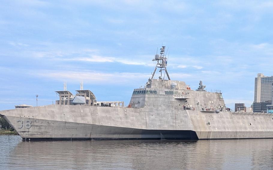 A gray-hulled military ship sails through a shipyard with multi-story buildings in the background.