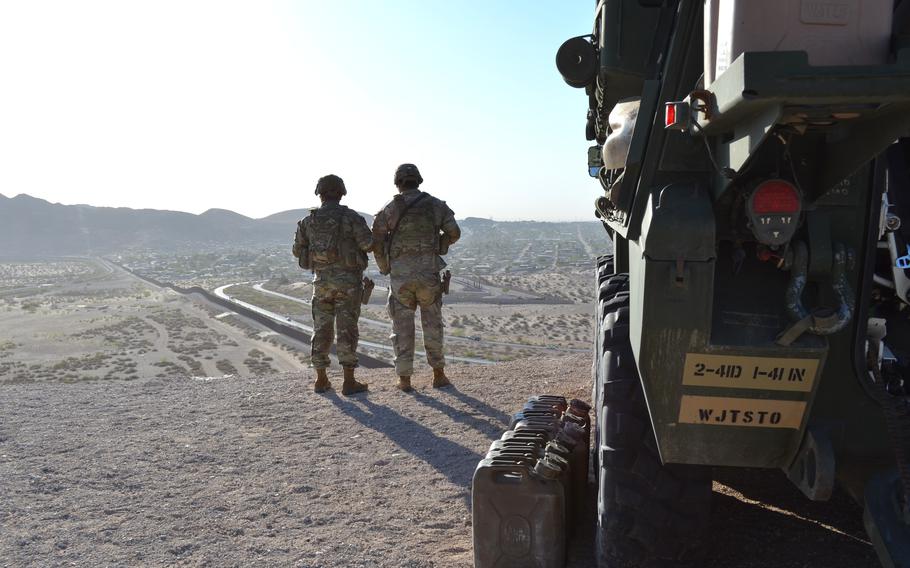 Two soldiers seen from the back as they look out at a vast empty area of desert. A military vehicle is behind them.