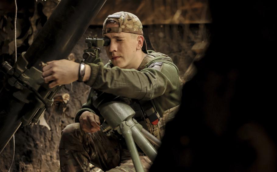 A soldier in camouflage combat gear kneels down next to a mortar and looks into a sight scope.