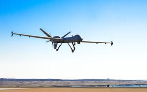 A military drone flies low over a sparse landscape with a clear blue sky and the horizon in the background.
