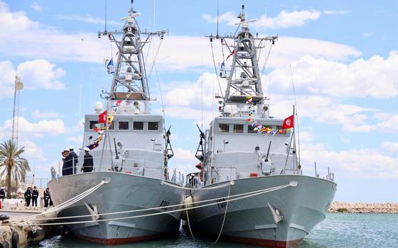 Two gray military patrol boats are docked side by side at a pier, adorned with colorful signal flags and the red Tunisian national flag. Several naval personnel stand on the deck of the left vessel, and a group of people is visible onshore.