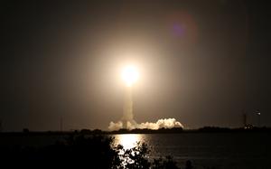 A bright light in the middle of a dark sky caps off a large plume of smoke.