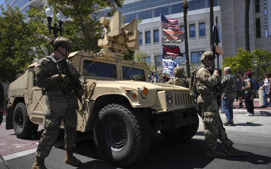 Two armed men stan on either side of an armored vehicle.