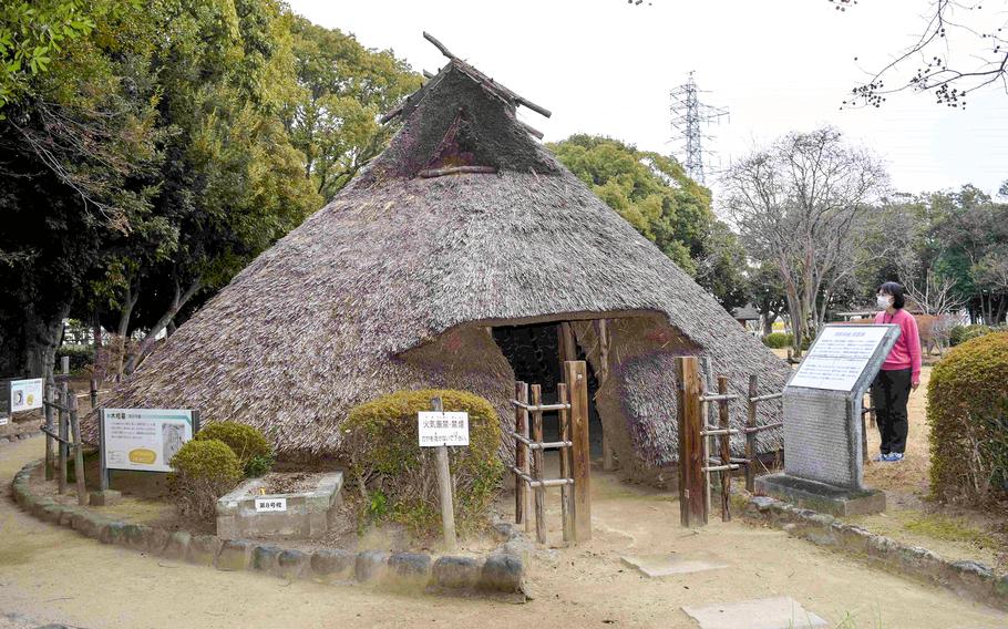 An exhibit of a replica of an ancient dwelling made of wood and straw is surrounded by a dirt path with plaques and markers displaying historical information.