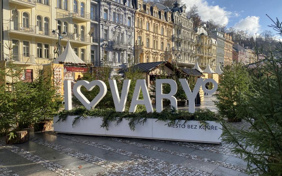 Winter holiday greenery decorates the pedestrian zone in front of the Mill Colonnade in Karlovy Vary, Czech Republic, with the resort town's pastel-colored buildings serving as a backdrop.