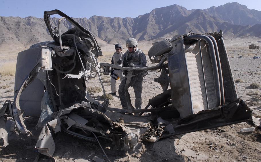 Two service members stand between two halves of a vehicle torn apart by an explosion.