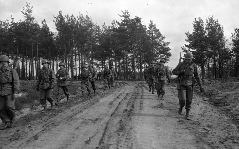 A black and white photo of soldiers walking through a mud/dirt road in a forested area.
