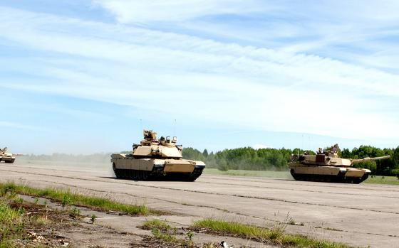 Multiple M1 Abrams tanks conduct training exercises. The tanks are positioned across a wide paved area with dust visible around them.