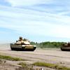 Multiple M1 Abrams tanks conduct training exercises. The tanks are positioned across a wide paved area with dust visible around them.