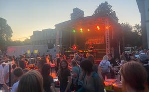 A crowd of people illuminated by the lights of a concert stage amid a setting sun.