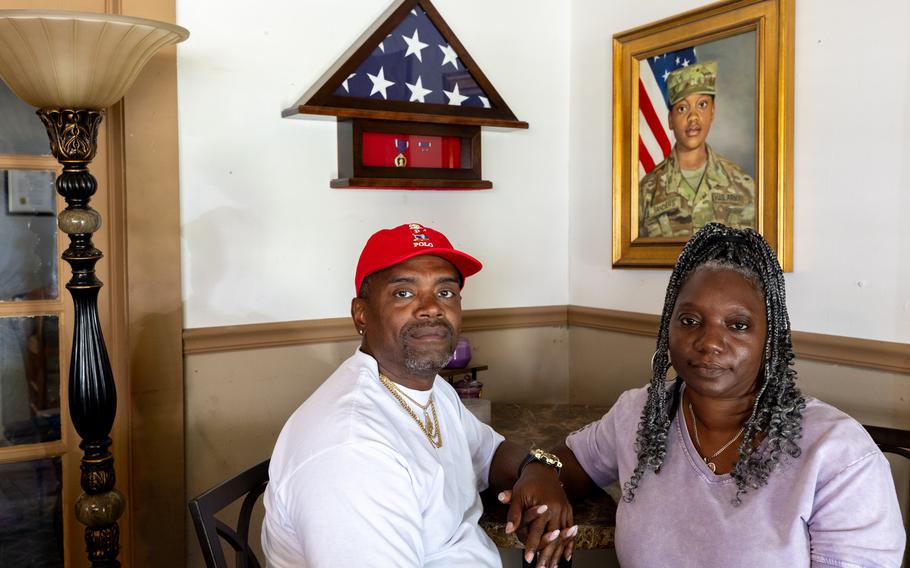 A man and a woman hold hands while seated at a table below a portrait of their late daughter in uniform.