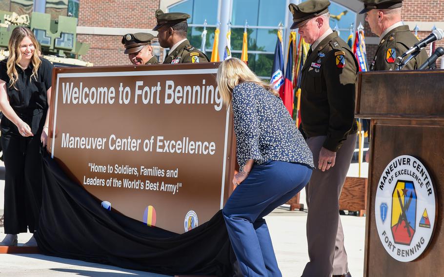 Two women remove a black drape to reveal a brown “Welcome to Fort Benning” sign. Some soldiers stand and watch.