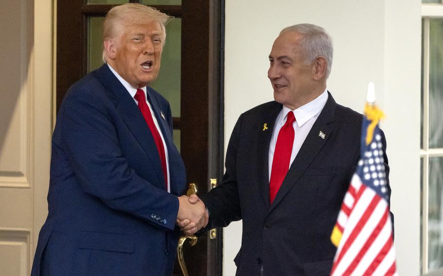 Donald Trump and Benjamin Netanyahu shake hands while standing in front of a door outside the White House, with a small U.S. flag in the foreground.