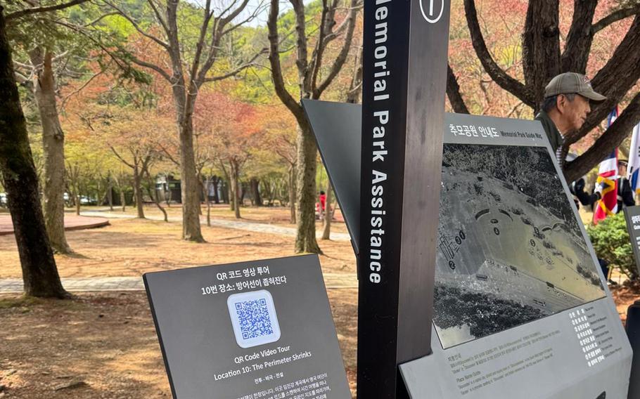 An informational sign for “Memorial Park Assistance” is shown in a park setting with autumn foliage, featuring Korean and English text, a QR code for video tours, and a person in a cap reading the display among trees with colorful fall leaves.