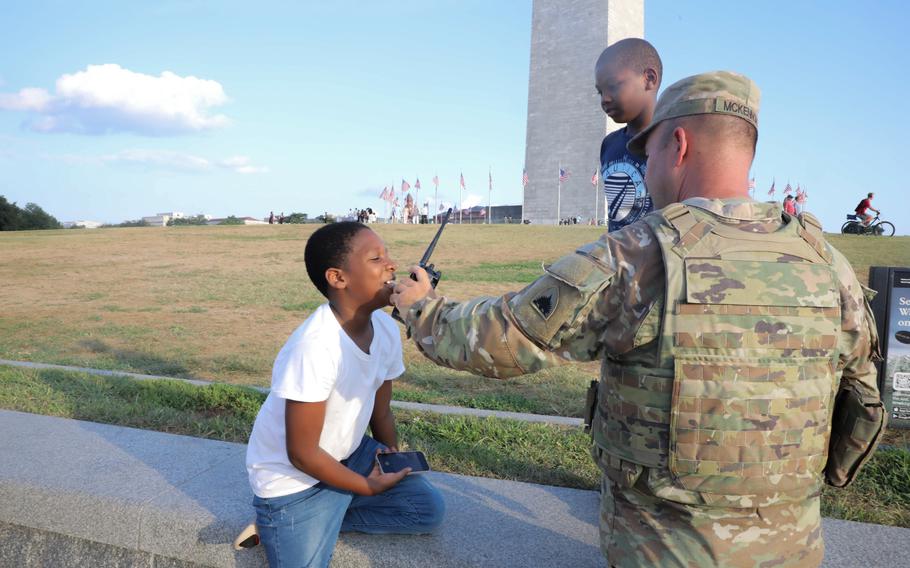 Two little boys next to a soldier with the Washington Monument visible in the background nearby; the soldier is holding a radio to one boy’s face as the boy smiles.