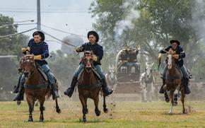 Soldiers in western-style blue cavalry uniforms ride horses at full gallop.