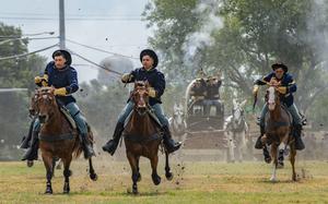 Soldiers in western-style blue cavalry uniforms ride horses at full gallop.