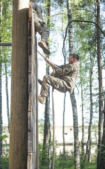 A vertical photo of soldiers climbing up a rope wall in a forest.