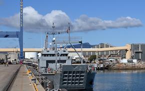 A gray military naval vessel is moored at a pier. The ship has a distinctive grid-like pattern on its stern and various communication equipment and masts visible on its superstructure. 