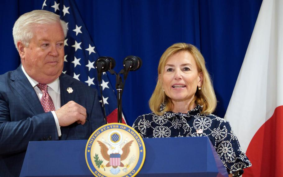A woman in a patterned navy dress stands at a podium with the United States Embassy seal, speaking while a man in a suit stands beside her. Behind them are the U.S. and Japanese flags.