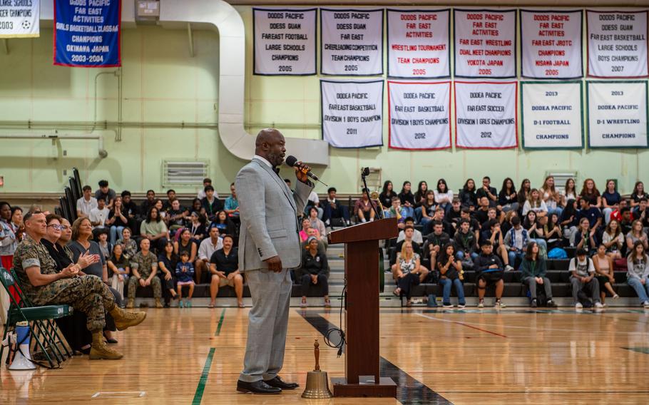 A man in a suit speaks behind a podium in a gymanasium full of students.