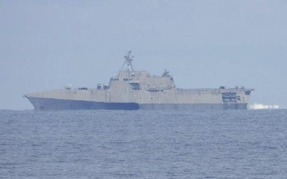 A naval warship, seen from a distance, sails in open water with the hazy horizon in the background.
